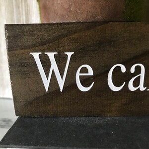 May include: A dark brown wooden sign with white lettering that reads "We can".