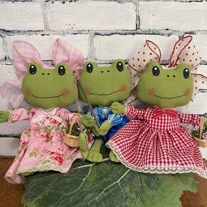 May include: Three plush frog dolls with green bodies, rosy cheeks, and large eyes. Each frog wears a different outfit: a pink floral dress, a blue plaid shirt and pants, and a red gingham dress. They sit on a large green leaf, with a white brick background.