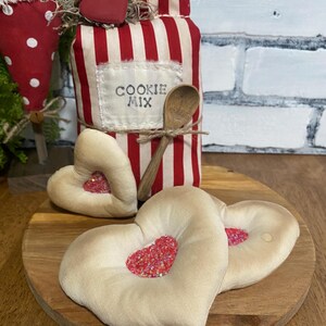 May include: A decorative jar with red and white stripes, labeled "COOKIE MIX", is adorned with a wooden spoon and a red heart. Two heart-shaped pillows with red glitter centers rest on a wooden board, creating a charming, rustic aesthetic.