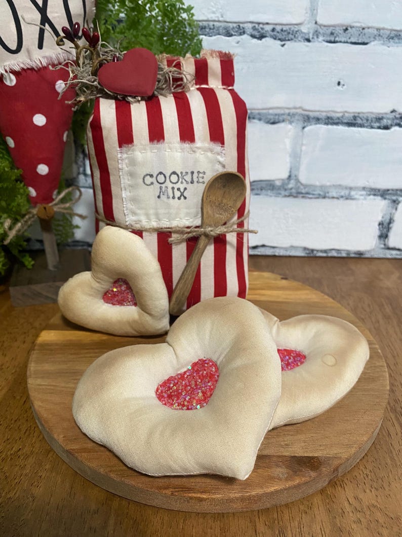 May include: A decorative jar with red and white stripes, labeled "COOKIE MIX", is adorned with a wooden spoon and a red heart. Two heart-shaped pillows with red glitter centers rest on a wooden board, creating a charming, rustic aesthetic.