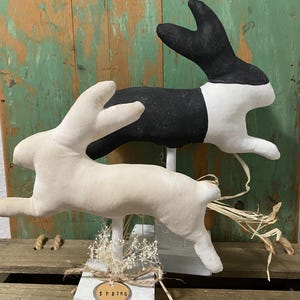 May include: Two fabric bunny figurines, one white and one black and white, standing on white wooden bases with a brown wooden sign that says "SPRING".