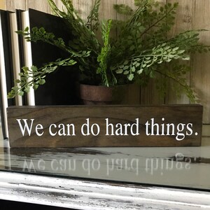 May include: A wooden sign with the text "We can do hard things." in white lettering. The sign is brown and has a rustic look.