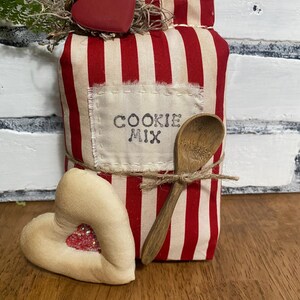 May include: A decorative fabric cookie mix container with red and white stripes, a wooden spoon, and a heart-shaped accent. The container has the words "COOKIE MIX" printed on a sewn-on label. A second heart-shaped accent is also present.
