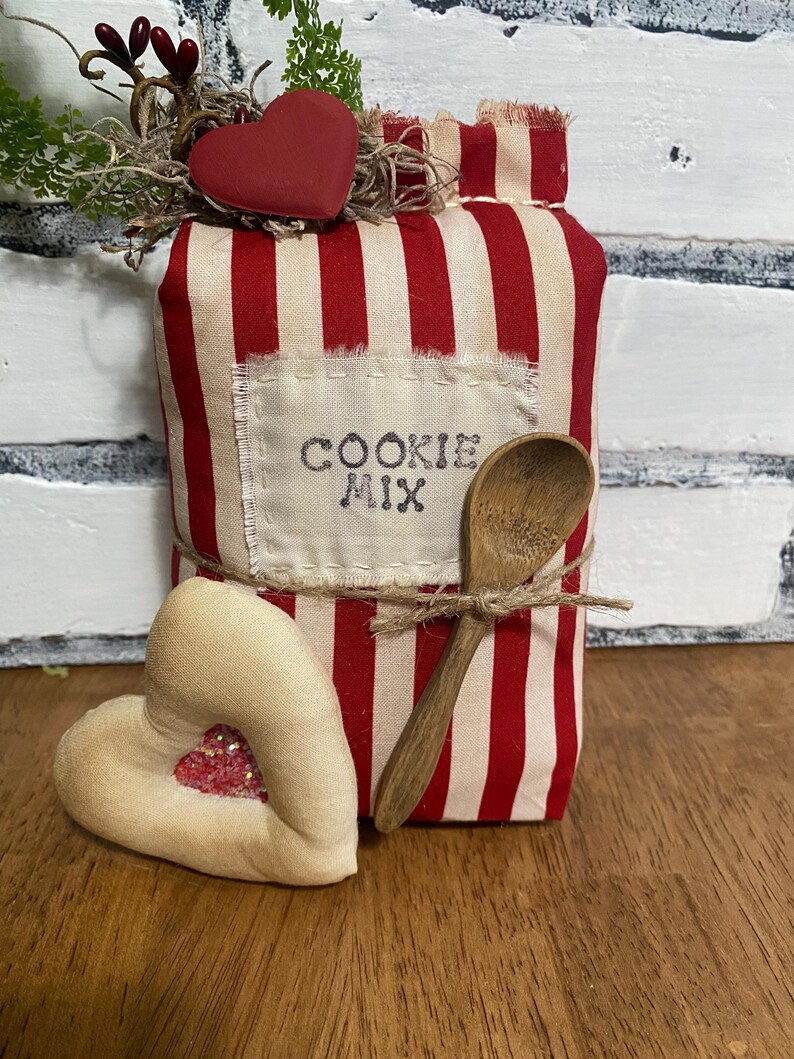 May include: A decorative fabric cookie mix container with red and white stripes, a wooden spoon, and a heart-shaped accent. The container has the words "COOKIE MIX" printed on a sewn-on label. A second heart-shaped accent is also present.