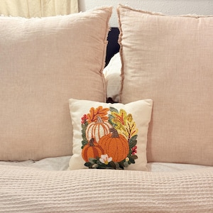 May include: A small square cream-colored throw pillow featuring a textured punch needle embroidery design of orange and white pumpkins surrounded by colorful autumn leaves, green foliage, and small red berries.