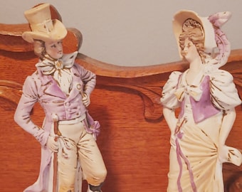 Puede incluir: Un par de figuritas de porcelana que representan a un hombre y una mujer con ropa vintage. El hombre lleva un sombrero de copa, una chaqueta morada y pantalones blancos. La mujer lleva un vestido blanco con un chaleco morado y un sombrero blanco con una pluma. Ambas figuritas están de pie sobre una base con un diseño floral.