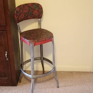 May include: Vintage metal bar stool with a floral patterned seat and backrest. The seat is trimmed in red, and the metal frame has a silver finish. The stool has a footrest and four legs.