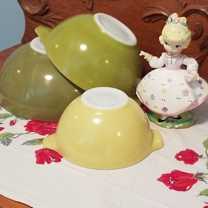 May include: Three vintage green and yellow mixing bowls stacked on a white tablecloth with a red floral pattern. A porcelain doll with a white dress and floral accents is in the background.