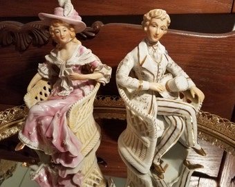 Puede incluir: Un par de figuritas de porcelana de un hombre y una mujer con atuendos del siglo XVIII, cada uno sentado en una silla de mimbre. La mujer lleva un vestido rosa y blanco con un sombrero blanco y el hombre lleva un traje blanco y dorado con un sombrero negro.