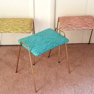 May include: Three gold-toned metal stools with fabric seats. The seats are in different colors and patterns: yellow with black stripes, pink with black stripes, and teal with black stripes.