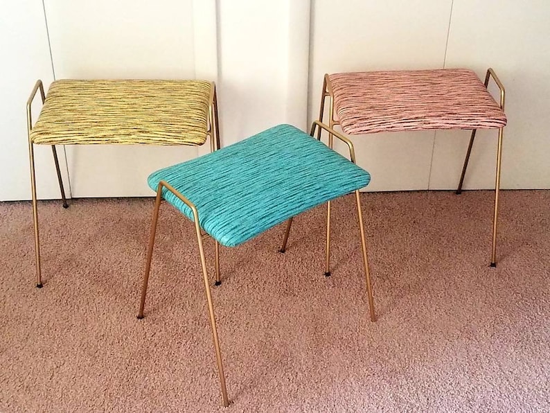 May include: Three gold-toned metal stools with fabric seats. The seats are in different colors and patterns: yellow with black stripes, pink with black stripes, and teal with black stripes.