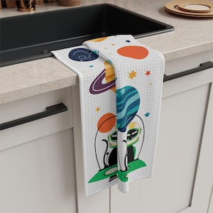 Op de afbeelding: Witte keukendoek met een kleurrijk ruimtethema-ontwerp. De handdoek toont planeten, sterren en twee zwarte buitenaardse figuren in ruimtehelmen. De handdoek hangt over een zwarte gootsteen.