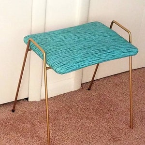 May include: A teal upholstered stool with a gold metal frame. The stool has a simple design with four legs and a single bar across the top.