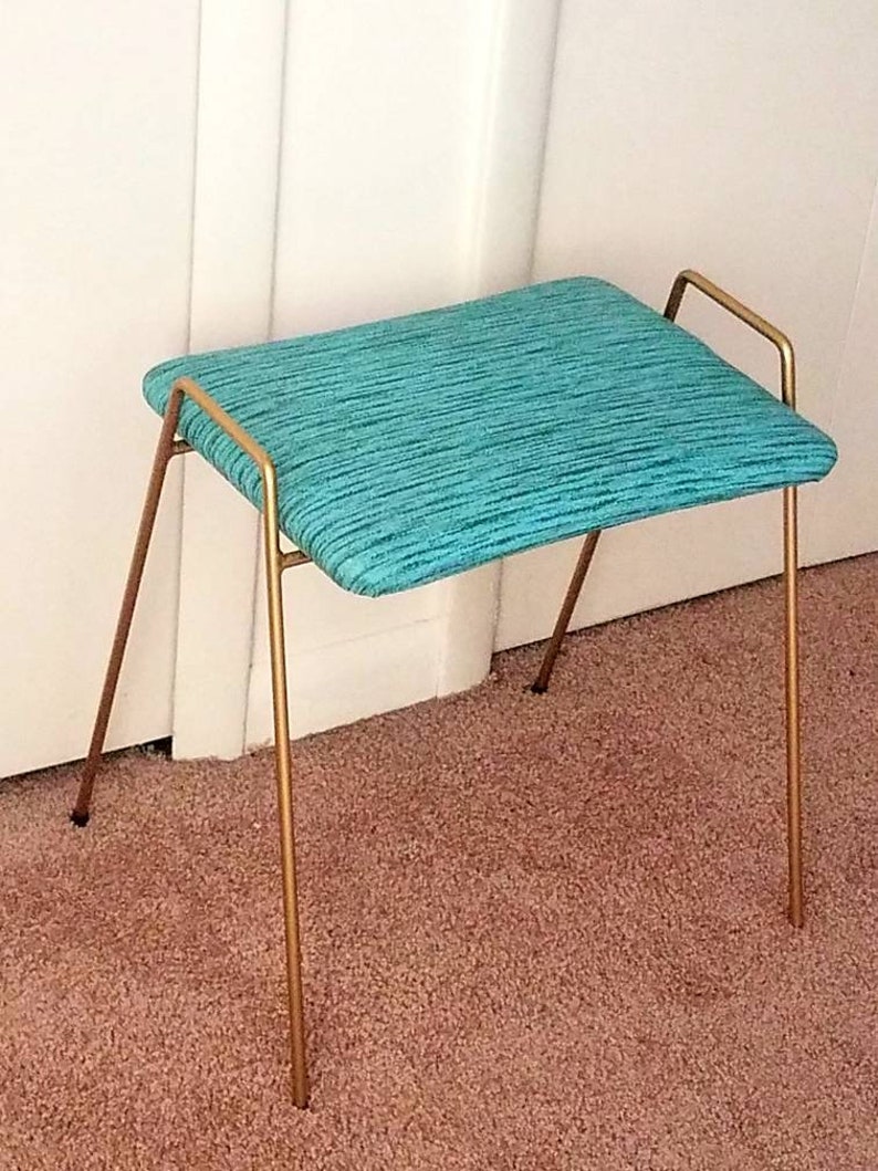 May include: A teal upholstered stool with a gold metal frame. The stool has a simple design with four legs and a single bar across the top.