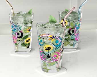 May include: Three clear glasses with colorful retro-style designs featuring a black cat, rockets, and planets. Each glass has a gold straw and is filled with ice and a green drink.