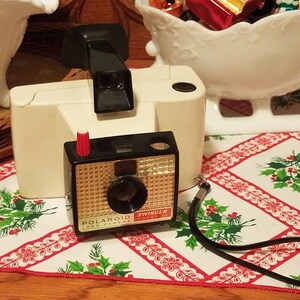 May include: A white Polaroid Land Camera Swinger model with a black lens and a red button. The camera is on a white and red tablecloth with a green holly pattern.