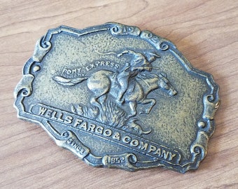 May include: A gold-toned belt buckle featuring a Wells Fargo & Company logo with a rider on horseback. The buckle has the text "Pony Express" and "Since 1852" around the edges.