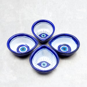 Può includere: Quattro piccole ciotole in ceramica bianca con bordi blu e un design di occhio turco blu. Le ciotole sono disposte a forma di fiore.