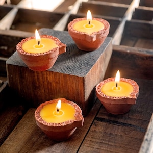 以下が含まれることがあります： 黄色い蜜蝋のろうそくが燃えている、小さなテラコッタ製のろうそく立てが4つ。ろうそく立ては木製の表面に置かれています。