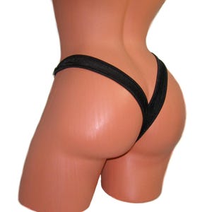 Puede incluir: Traje de baño negro estilo tanga con diseño en forma de V. La tela brillante crea un efecto metálico. La imagen se centra en la parte trasera del traje de baño, destacando el corte y el estilo.