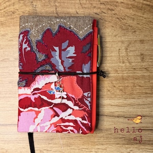 May include: A handmade journal cover with a floral pattern in shades of red, pink, and gray. The cover has a brown top section with white stitching and a black elastic closure. A dragonfly charm hangs from the elastic. A red pen is visible.
