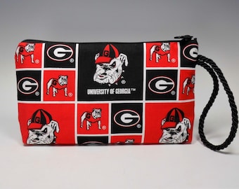 May include: A rectangular zippered pouch with a black wrist strap. The pouch features a repeating pattern of red and black squares with the University of Georgia bulldog logo and the letter "G". The text "UNIVERSITY OF GEORGIA" is also visible.