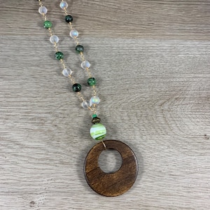 May include: A necklace featuring a gold-tone chain strung with alternating translucent white beads and dark green speckled beads. A single green and white striped bead sits above a large, circular, dark brown wooden pendant with a center cutout.