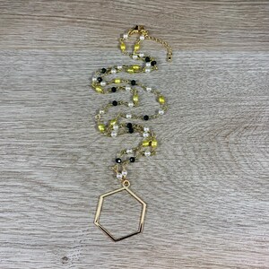 May include: Gold-toned necklace with a hexagonal pendant. The chain features a mix of white, black, and yellow beads. The necklace is displayed on a wooden surface.
