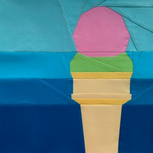 Könnte beinhalten: Ein farbenfroher Quiltblock mit einem Eiswaffel-Design. Das Eis hat eine rosa Kugel, eine grüne Schicht und eine gelbe Schicht, alles auf einem beigen Kegel. Der Hintergrund ist eine Mischung aus Blautönen.
