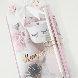 Könnte beinhalten: Ein rosa-weißes Notizbuch mit dem Wort "Glamorous" in goldener Schrift. Das Notizbuch hat ein Blumenmuster und enthält einen weißen Bleistift, ein rosa Gummiband und eine goldene Büroklammer. Eine weiße Tasse mit Wimpern und eine Uhr sind ebenfalls sichtbar.