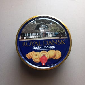 May include: A round blue metal tin of Royal Dansk Butter Cookies. The lid features a picture of a traditional house, the text Royal Dansk Butter Cookies, and an image of various cookies. The tin is labeled with a net weight of 454g.