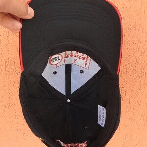 May include: A black baseball cap with a red trim on the brim. The interior features a white crown lining with a red logo patch reading The Rental Guys and a small black tag. A white care label is attached to the inner black fabric panel.