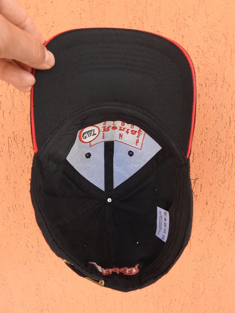 May include: A black baseball cap with a red trim on the brim. The interior features a white crown lining with a red logo patch reading The Rental Guys and a small black tag. A white care label is attached to the inner black fabric panel.