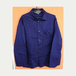 May include: A long-sleeved, solid dark blue button-down jacket featuring a standard collar, a single chest pocket on the left side, and five dark buttons running down the center front. The garment is displayed on a black plastic hanger.