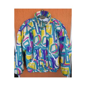 Peut inclure: Une veste de ski vintage avec un imprimé abstrait coloré dans les tons de bleu, jaune, rose et blanc. La veste a une fermeture éclair et une capuche.