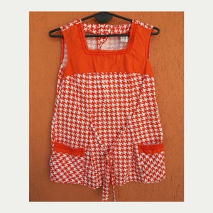 May include: Orange and white houndstooth print sleeveless top with a square neckline and two front pockets. The top has an orange yoke and trim, and a tie at the waist. The garment is hanging on a black hanger.