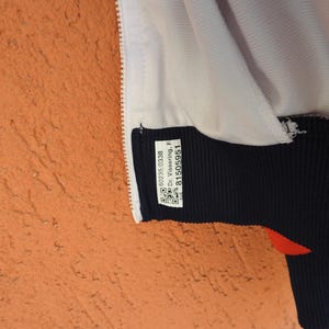 Puede incluir: Una chaqueta blanca con un dobladillo acanalado azul oscuro y una cremallera blanca. Una etiqueta blanca rectangular est&aacute; cosida en el dobladillo azul oscuro, con el texto 60235/0338, Dr. Vissering, F y 81505951, junto a dos c&oacute;digos QR.