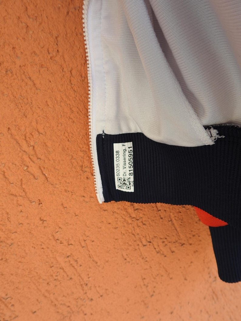 Puede incluir: Una chaqueta blanca con un dobladillo acanalado azul oscuro y una cremallera blanca. Una etiqueta blanca rectangular est&aacute; cosida en el dobladillo azul oscuro, con el texto 60235/0338, Dr. Vissering, F y 81505951, junto a dos c&oacute;digos QR.