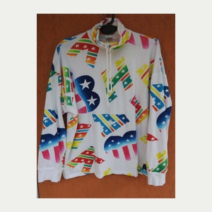 Puede incluir: Sudadera blanca de manga larga con un estampado colorido de estrellas y rayas. El estampado presenta una gama de colores arcoíris con rojo, naranja, amarillo, verde, azul y morado.