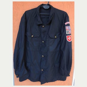 May include: A dark navy blue jacket with four front pockets and gold-coloured buttons. The jacket has three patches on the left sleeve, including a red cross and a crest. The jacket is hanging on a black hanger.