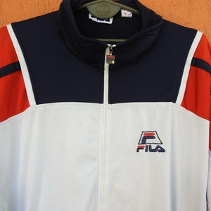 Puede incluir: Una chaqueta deportiva con cremallera que presenta un dise&ntilde;o de bloques de color en azul marino, blanco y rojo. La chaqueta tiene cuello alto, mangas largas y el logotipo de Fila bordado en azul marino y rojo en el pecho izquierdo.