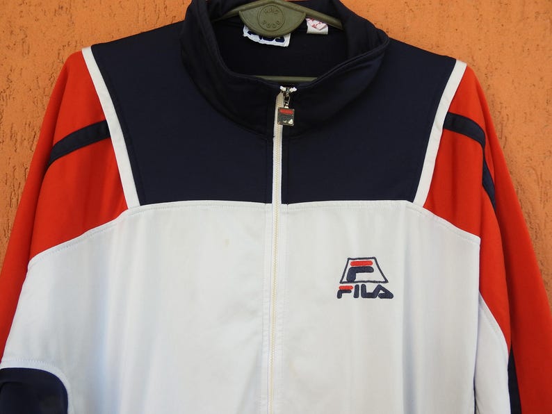 Puede incluir: Una chaqueta deportiva con cremallera que presenta un dise&ntilde;o de bloques de color en azul marino, blanco y rojo. La chaqueta tiene cuello alto, mangas largas y el logotipo de Fila bordado en azul marino y rojo en el pecho izquierdo.