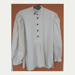 May include: A long-sleeved off-white shirt featuring a mandarin collar, a partial button-down front with five dark brown buttons, and pleated detailing on the chest. The garment has gathered shoulders and cuffs, with the brand name Moon visible.