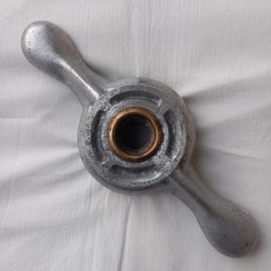Peut inclure: Une poignée en métal argenté avec un composant central circulaire et deux bras incurvés s'étendant vers l'extérieur. Le centre présente un anneau de couleur laiton autour d'un trou sombre. La poignée est probablement pour une vanne ou un mécanisme similaire.