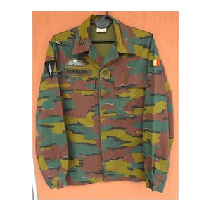 May include: A long-sleeve camouflage jacket featuring a button-down front, two chest pockets, and a collar. The jacket displays a parachute patch, a name tag reading VERHEUEN, a black triangular patch with a white sword, and a Belgian flag patch.