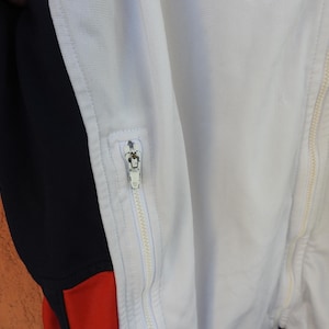 Puede incluir: Una chaqueta deportiva blanca con un panel lateral negro vertical y un acento rojo en la parte inferior. La prenda incluye una cremallera central de longitud completa y un bolsillo con cremallera m&aacute;s peque&ntilde;o en el frente, ambos con tiradores de metal plateado.