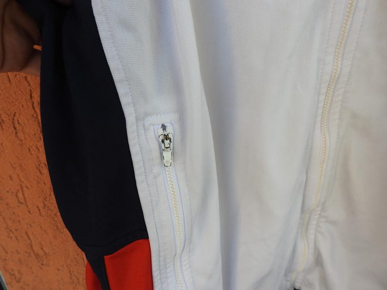 Puede incluir: Una chaqueta deportiva blanca con un panel lateral negro vertical y un acento rojo en la parte inferior. La prenda incluye una cremallera central de longitud completa y un bolsillo con cremallera m&aacute;s peque&ntilde;o en el frente, ambos con tiradores de metal plateado.