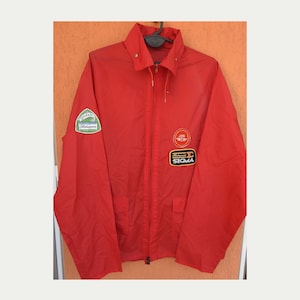May include: A bright red windbreaker jacket featuring a full-length front zipper, a pointed collar, and two front pockets. The jacket displays a green and white Shakespeare patch on the right sleeve and two patches on the left chest, including a circular red logo and a rectangular black patch with the text Sigma.