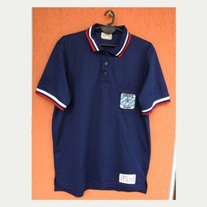 May include: A navy blue polo shirt with red and white trim on the collar and sleeves. The shirt has a pocket on the left chest with a patch that says "BBSV Umpire" and features a blue and white logo.