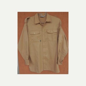Op de afbeelding: Een beige overhemd met knopen, twee borstzakken en lange mouwen. Het overhemd heeft een kraag en epauletten op de schouders. Een klein groen label is zichtbaar aan de voorkant. Het overhemd hangt aan een hanger.